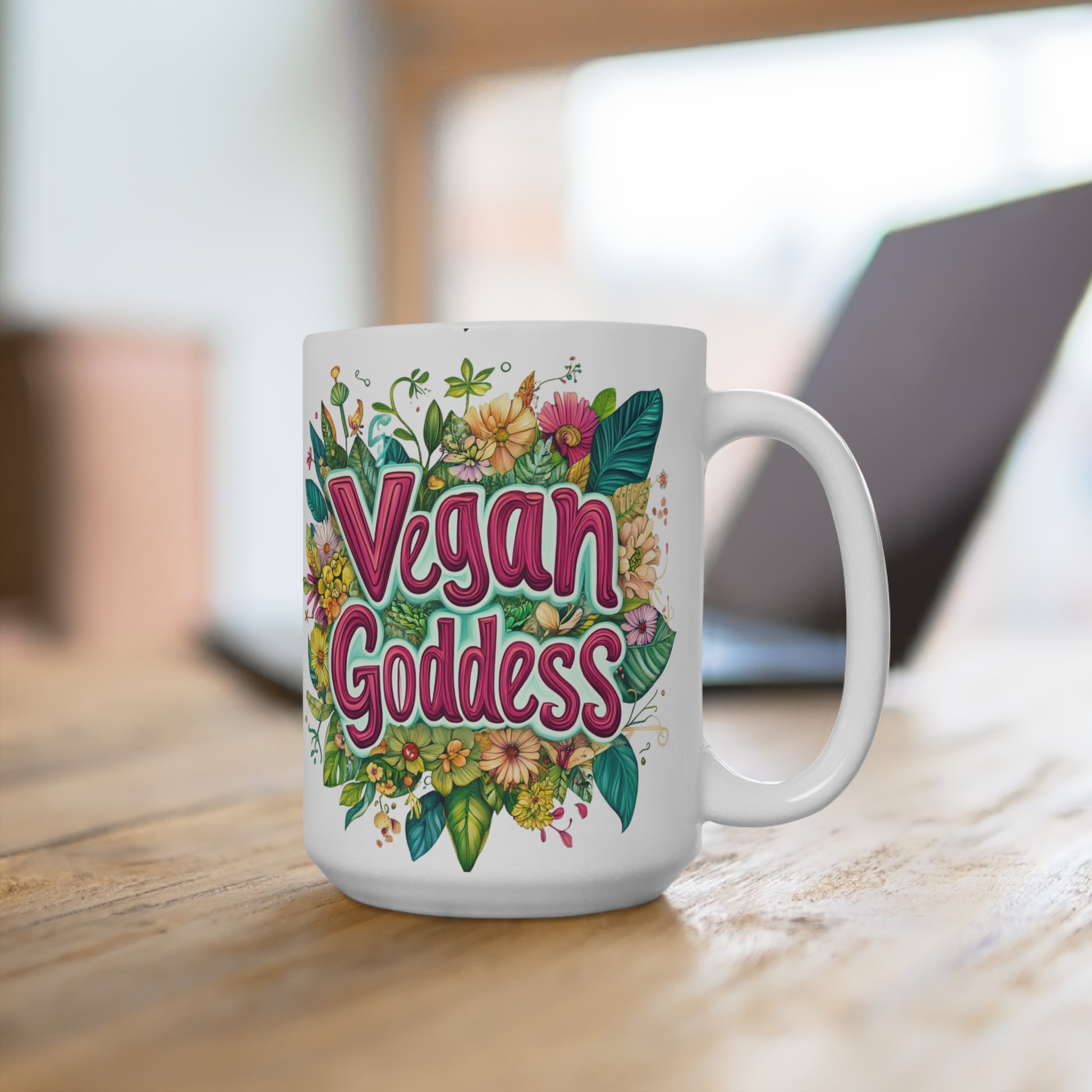 White ceramic vegan goddess mug featuring colorful floral artwork and bold pink lettering on a wooden desk with blurred laptop